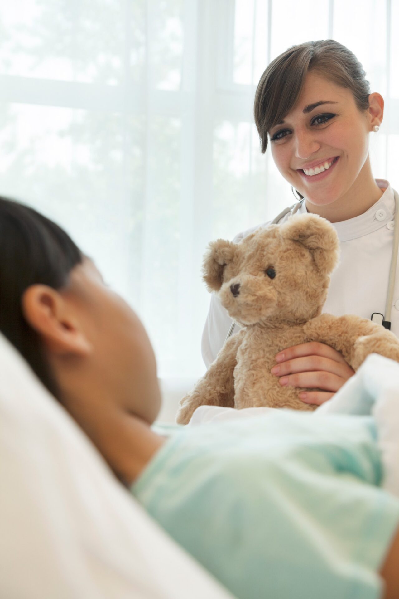 Smiling female doctor giving a teddy bear to a girl patient lying down on a hospital bed<br />
